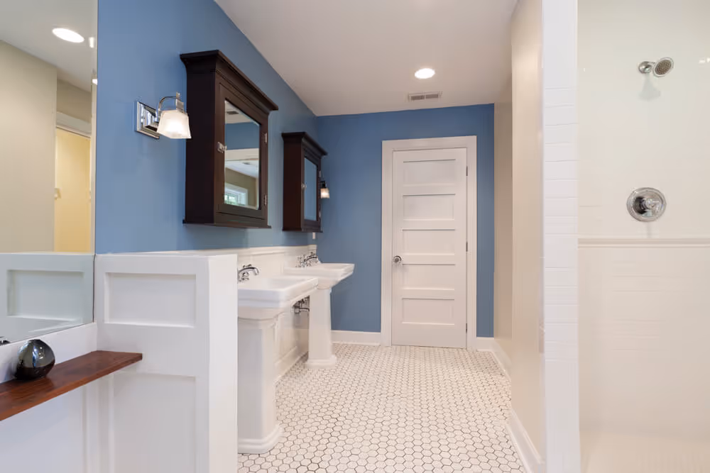 Modern bathroom with blue walls, white tile hexagon floor, two pedestal sinks with dark wooden medicine cabinets, and a white door.
