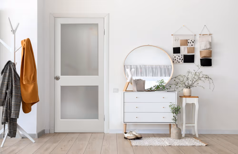 Minimalist entryway featuring a white door, coat rack with jackets, a dresser with a round mirror, and potted plants on wooden floor.