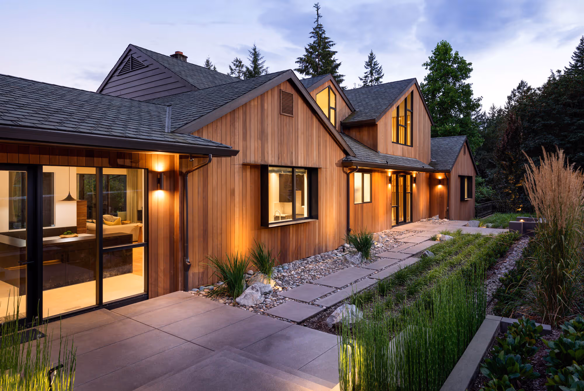 Contemporary wooden house exterior with warm interior lights, landscaped stone pathway, and greenery at dusk.