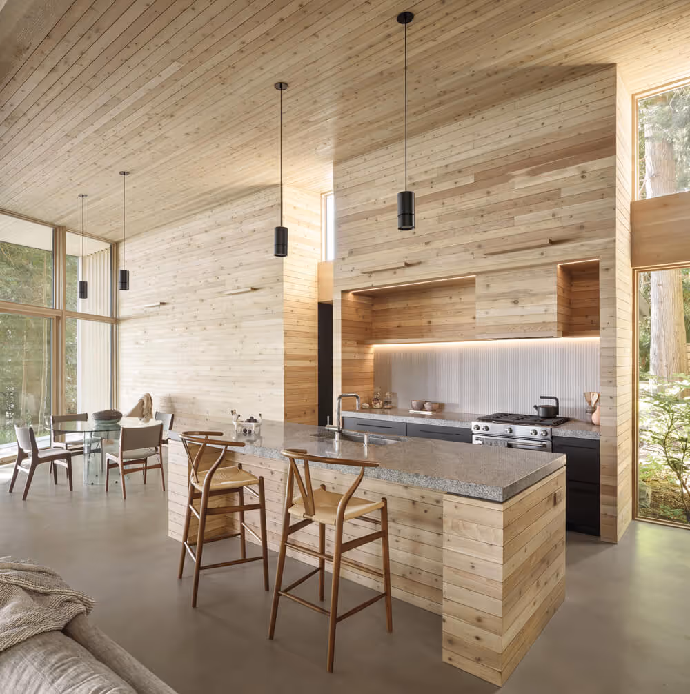 Modern kitchen with light wood walls and ceiling, gray concrete floor, stone countertop island with wooden stools, and large windows overlooking greenery.