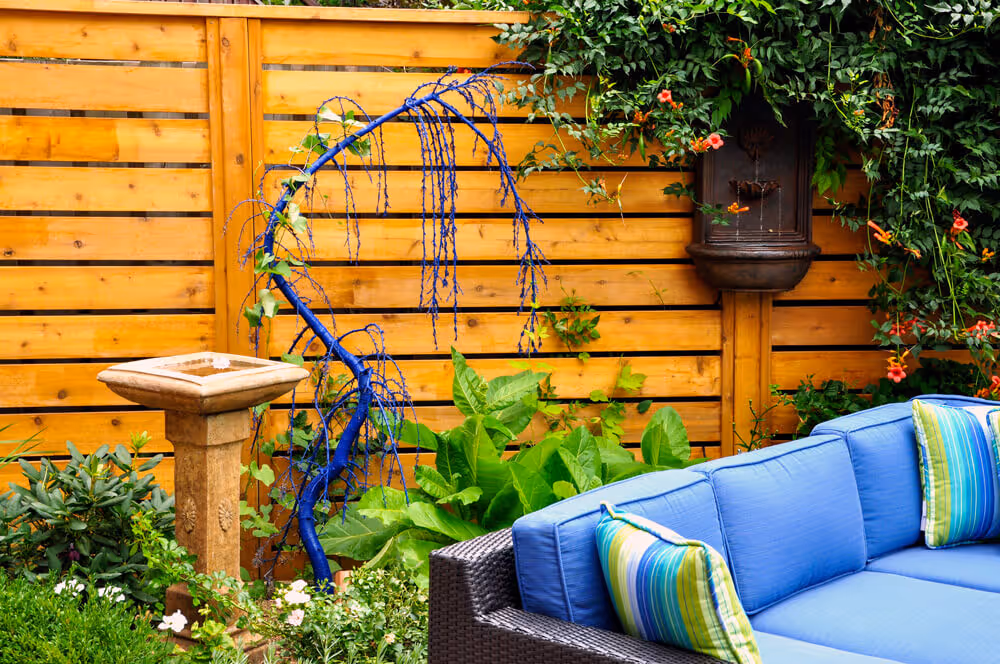 Outdoor patio area with a blue cushioned wicker sofa, green plants, a wooden birdbath, and a wooden fence background.
