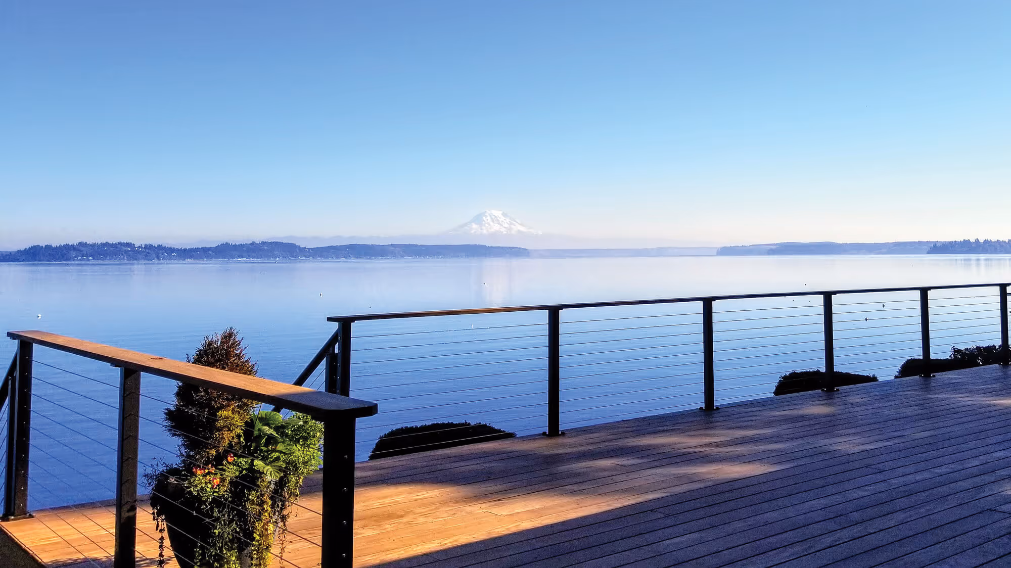 Wooden deck with cable railing overlooking a calm lake and distant snow-capped Mount Rainier under a clear blue sky.