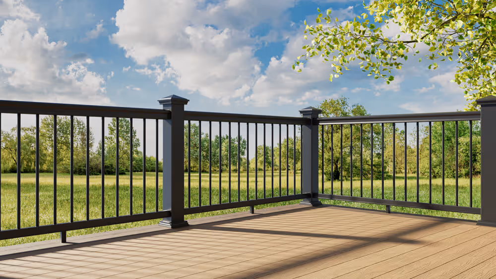Black metal railing on a wooden deck overlooking a green field with trees under a partly cloudy blue sky.