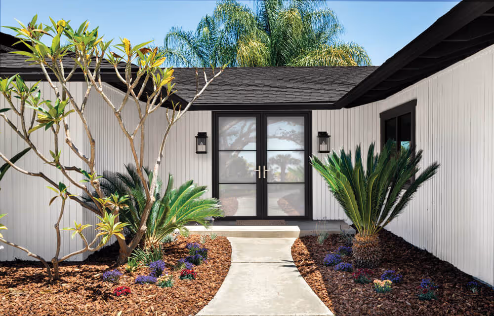 Modern house entrance with black framed glass double doors, white vertical siding, and landscaped garden with tropical plants and flowers.