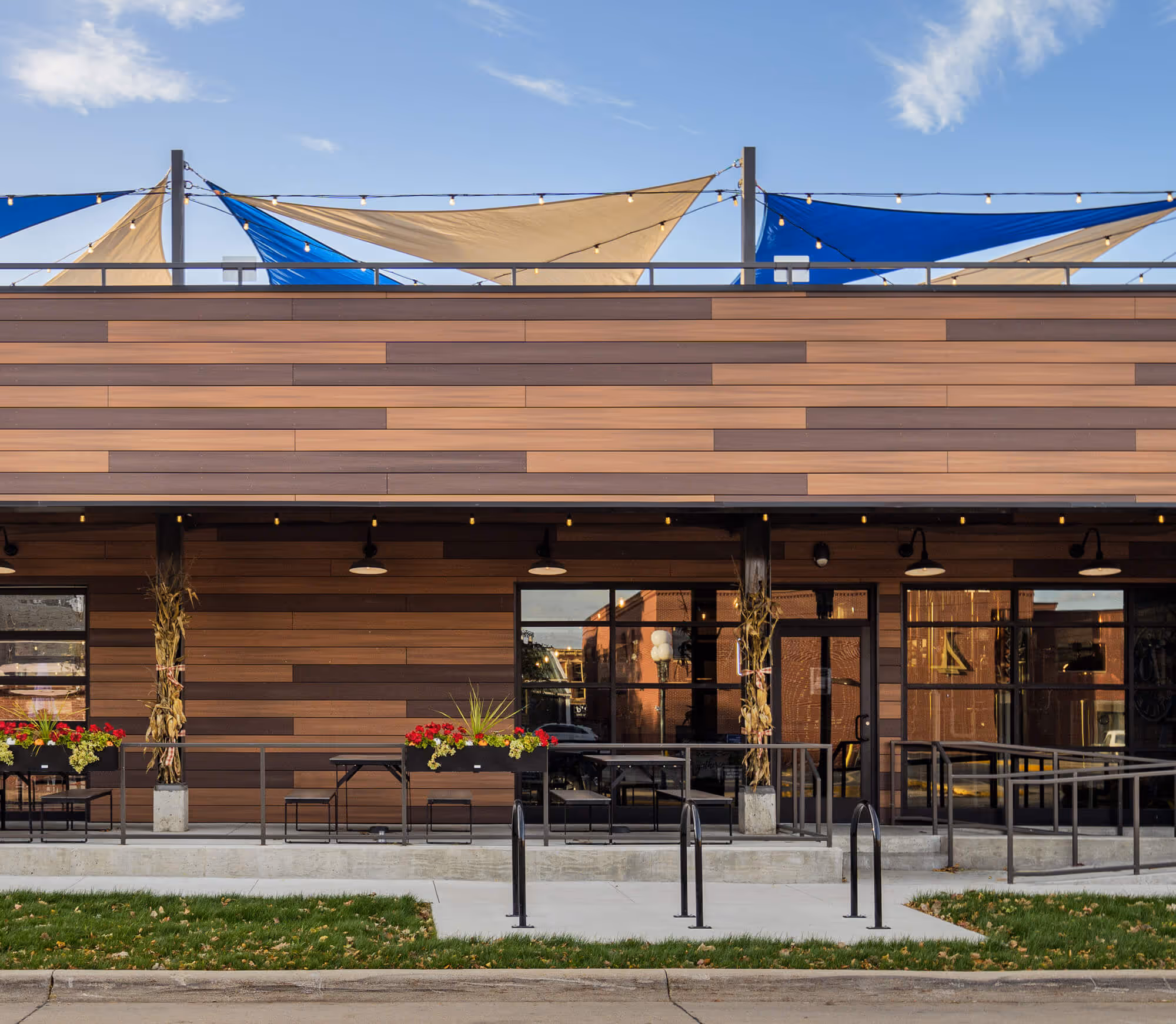 Modern building facade with wood paneling, outdoor seating with flower boxes, and blue and beige sun shades above.