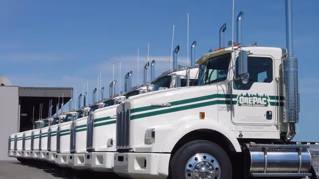 Line of white Kenworth trucks with green OrePac logos parked outside under clear blue sky.