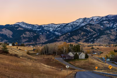 Rural Bozeman, MT, landscape with houses, winding road, and snow-capped mountains under a sunset sky.