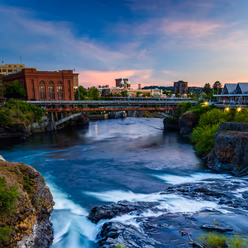 River flowing under a bridge with buildings and trees on both sides during sunset with a colorful sky.