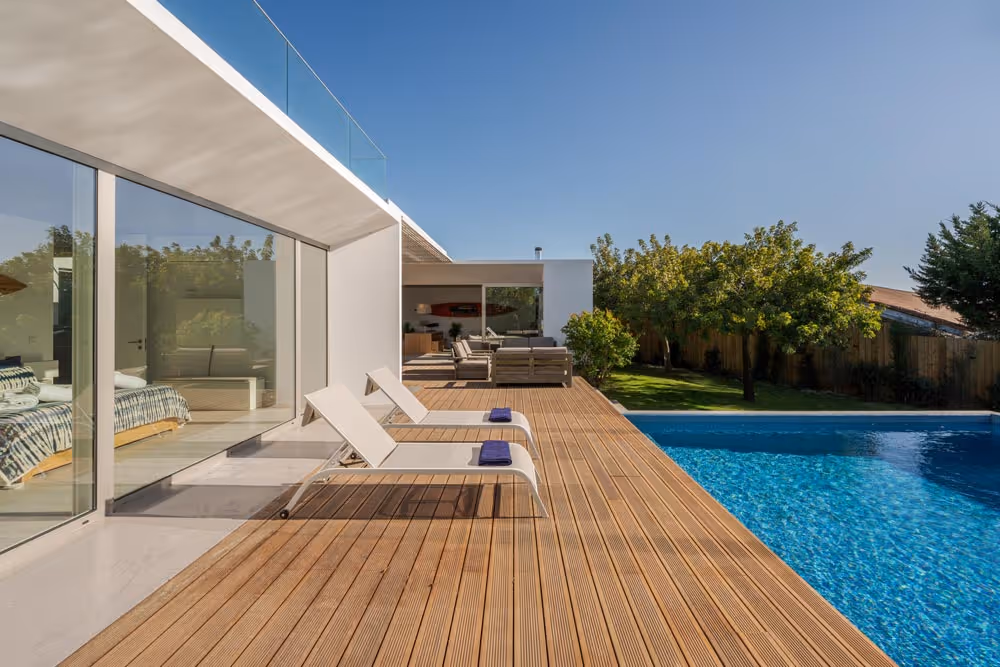 Modern house with a wooden deck featuring two white lounge chairs with towels, overlooking a clear blue swimming pool and a green yard with trees.