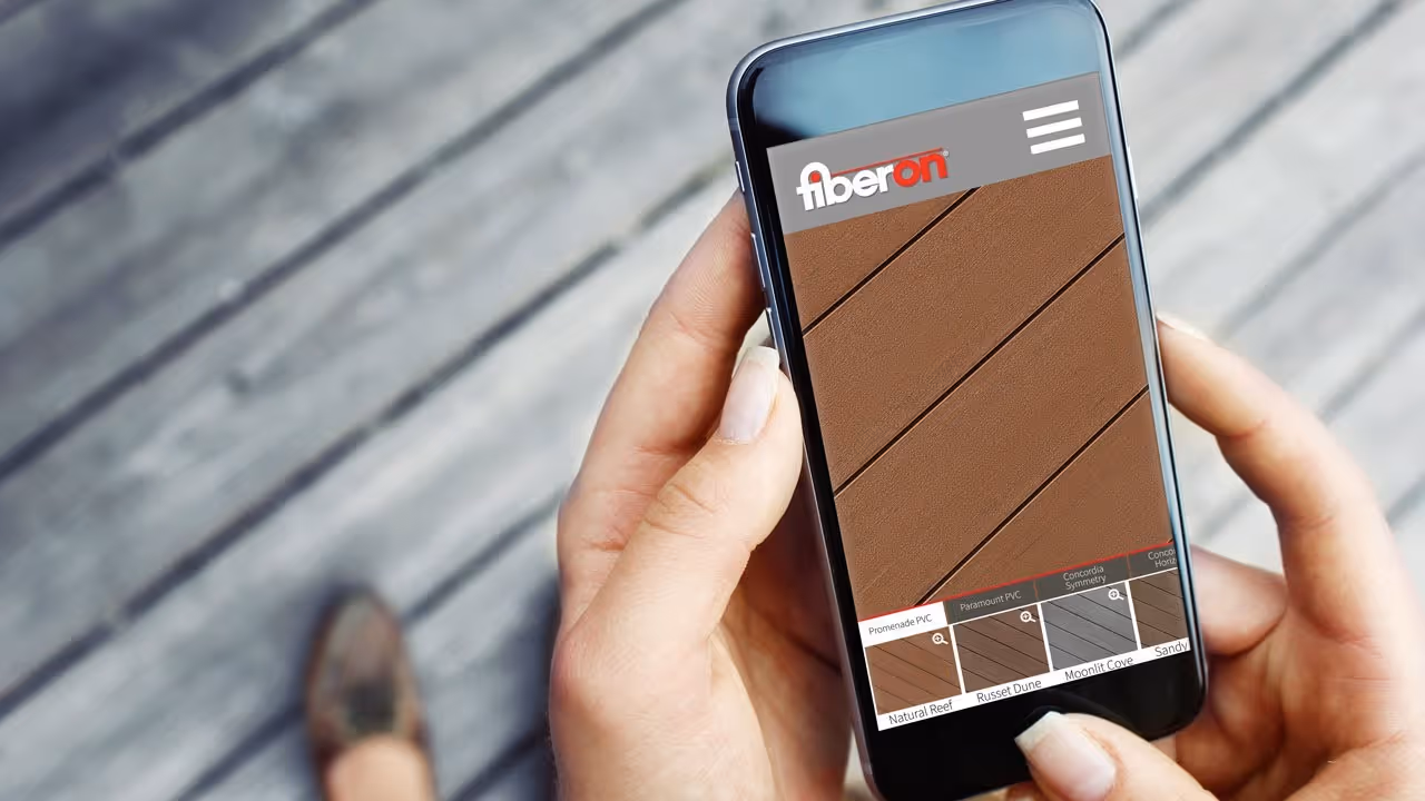 Person holding a smartphone displaying the Fiberon app interface with decking sample options and close-up textures.