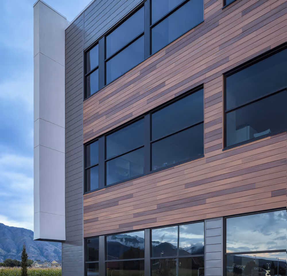 Modern building facade with mixed horizontal wooden and gray cladding panels and large reflective windows showing mountain and cloudy sky reflections.
