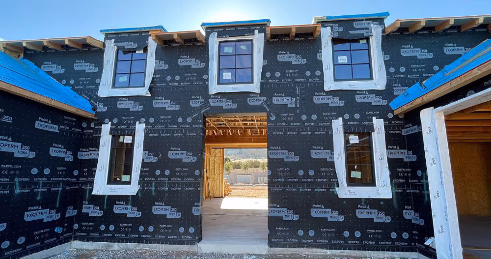 Front view of a house under construction with black weather-resistant wrap and multiple windows installed.