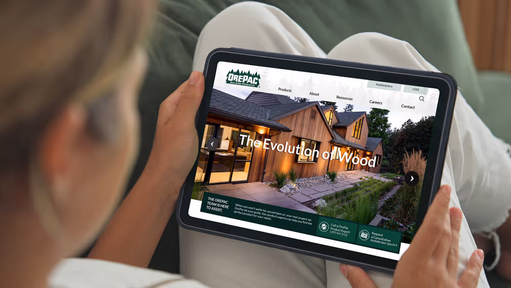 Person holding a tablet displaying OrePac building products website featuring a modern wooden house with the text 'The Evolution of Wood'.