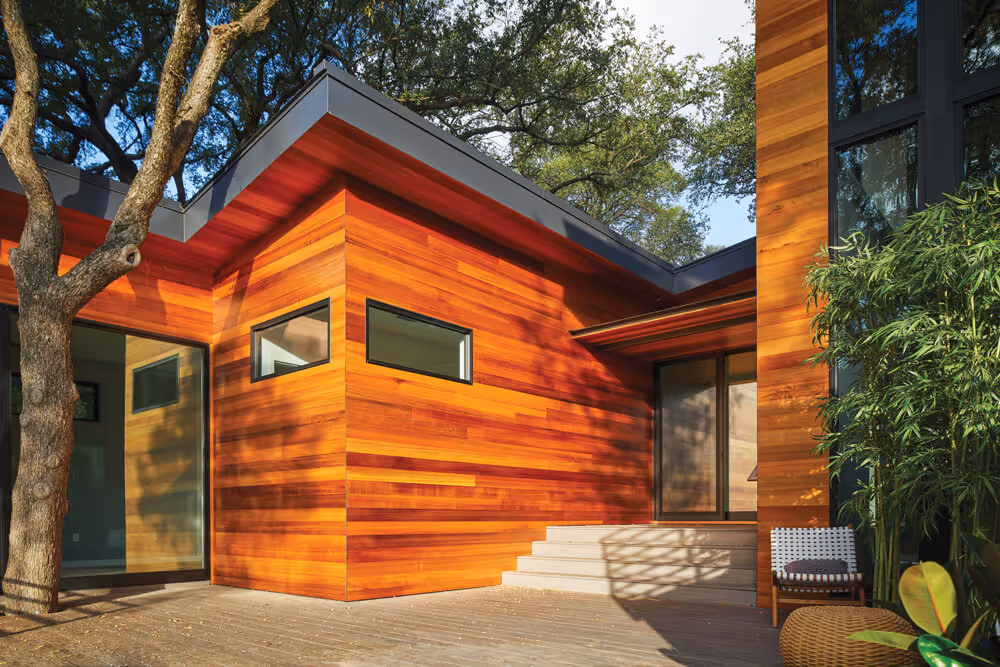 Modern house exterior with warm cedar wood siding, large windows, a deck, and surrounding trees.