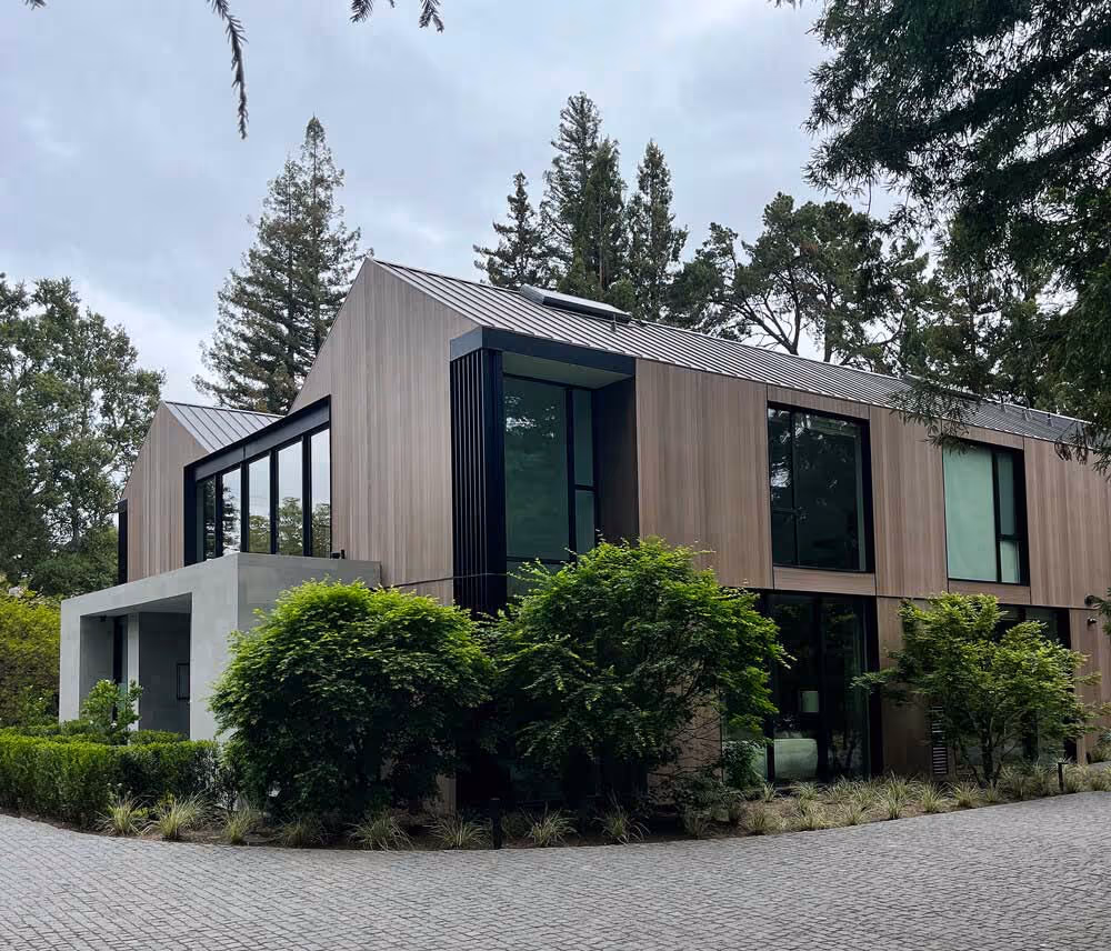 Modern two-story house with large windows, wood panel siding, and a gray metal roof surrounded by green trees and shrubs on a cobblestone driveway.