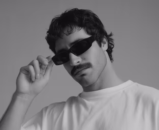 Man with curly hair and mustache wearing rectangular sunglasses and a white shirt, adjusting the sunglasses with his hand.