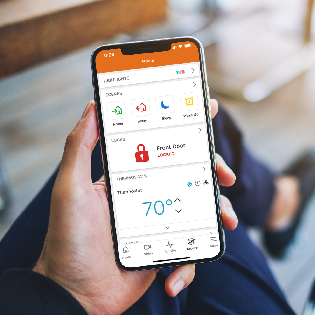 Hand holding smartphone showing home automation app with lock status and thermostat at 70 degrees.