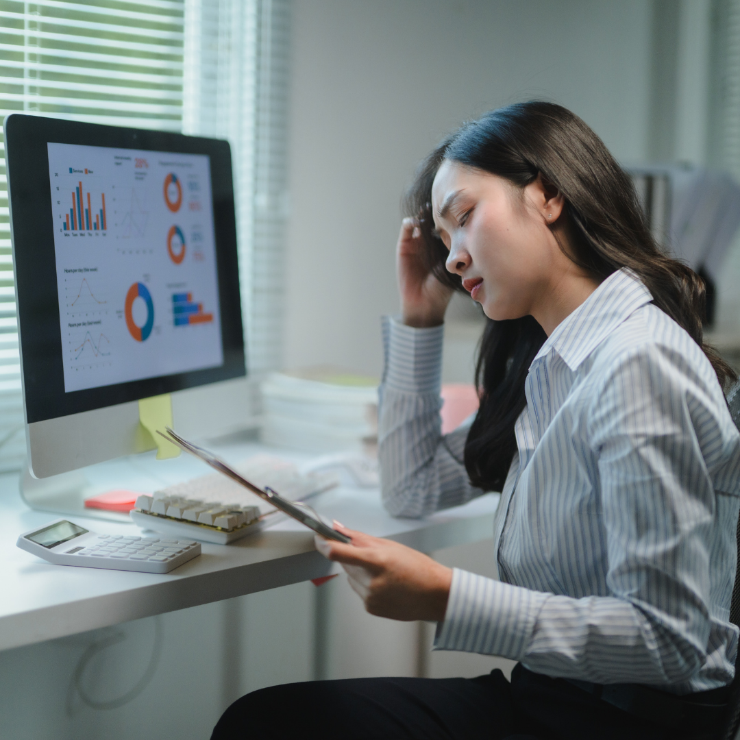 Stressed woman in striped shirt analyzing charts on paper and computer screen in office.