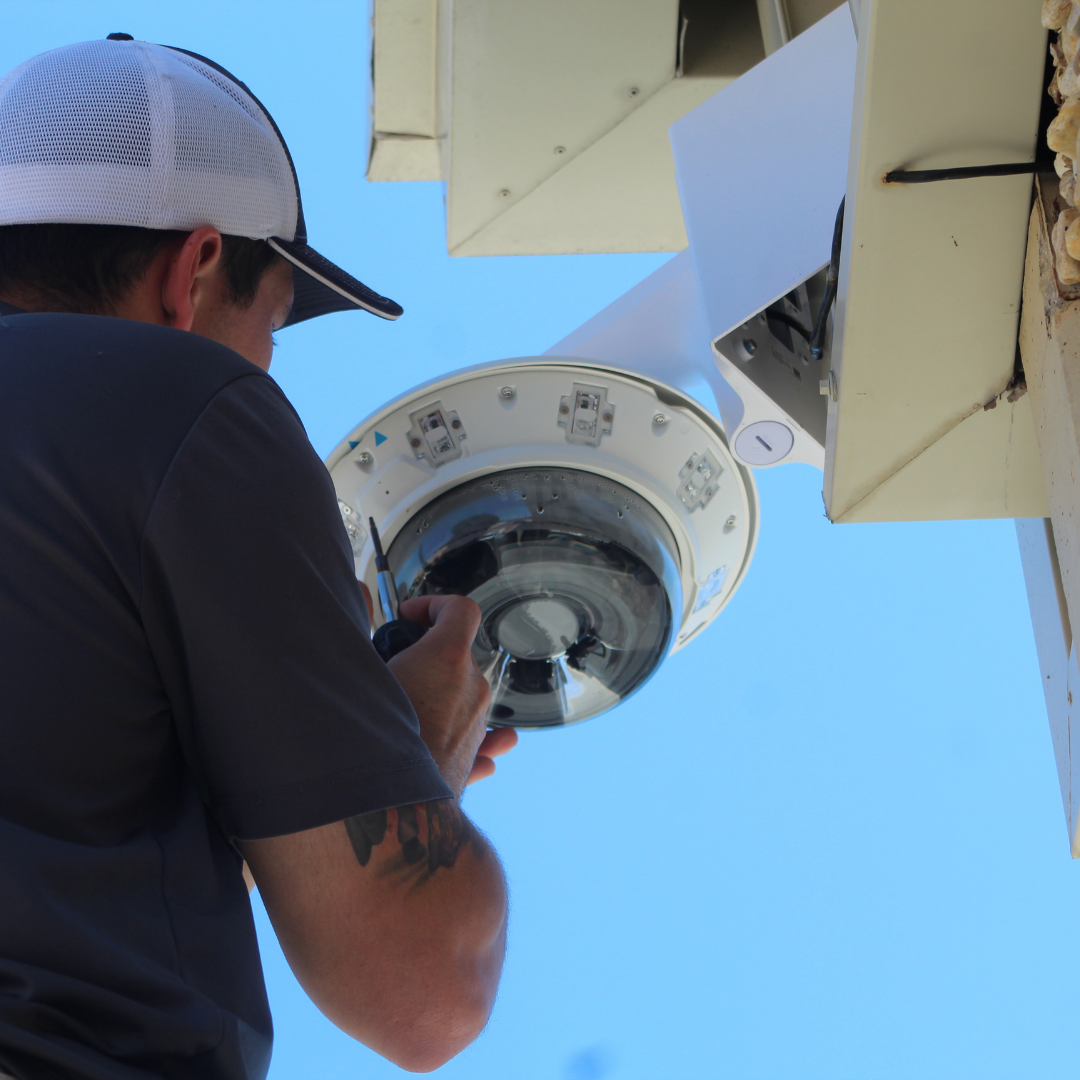 Technician wearing cap installs or repairs a dome security camera mounted on building exterior.