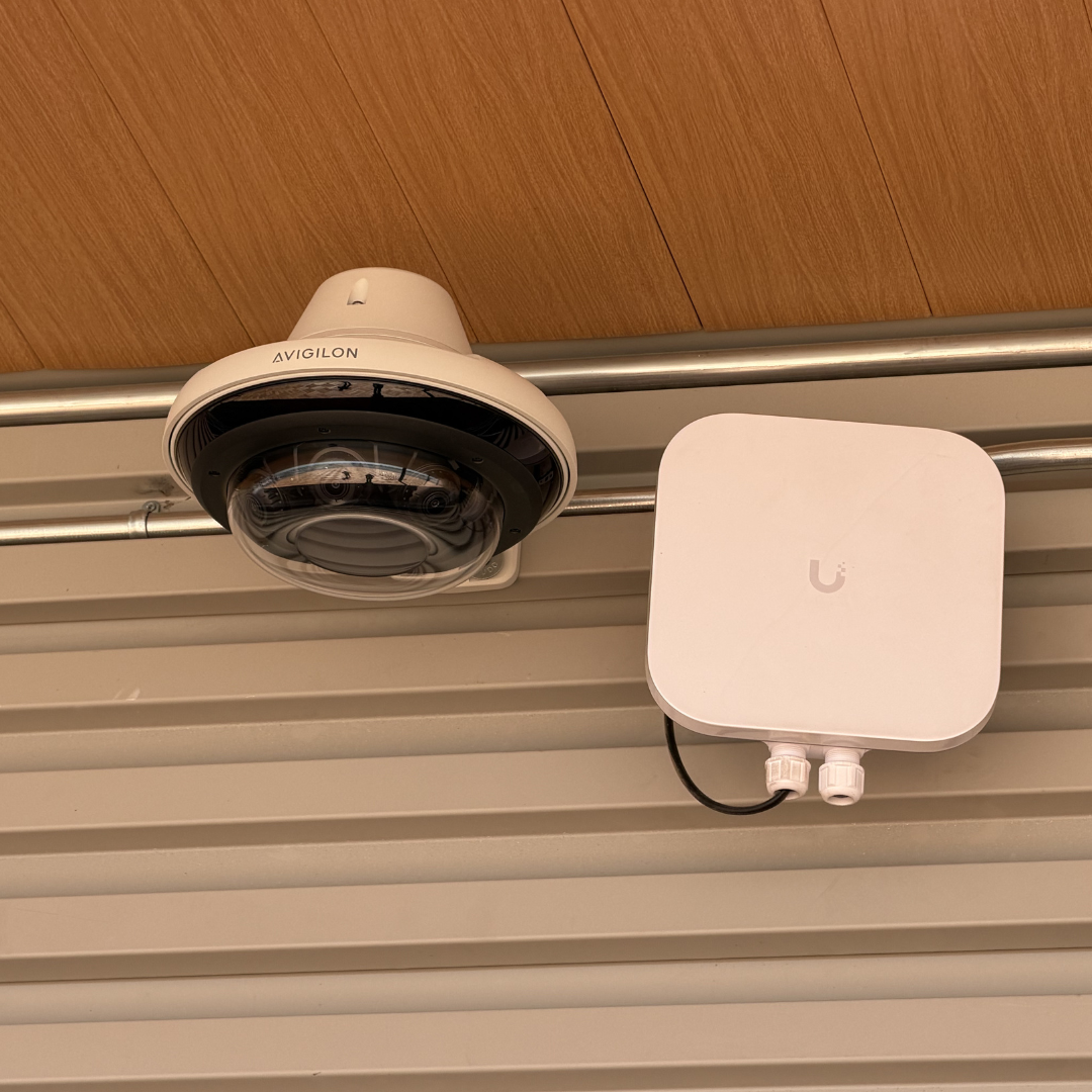 White dome security camera and a white network device mounted on a ceiling with wood paneling.