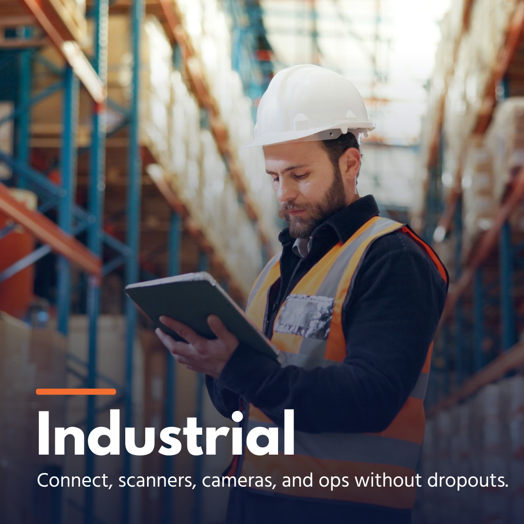 Man in white hard hat and orange vest using a tablet in a warehouse aisle.