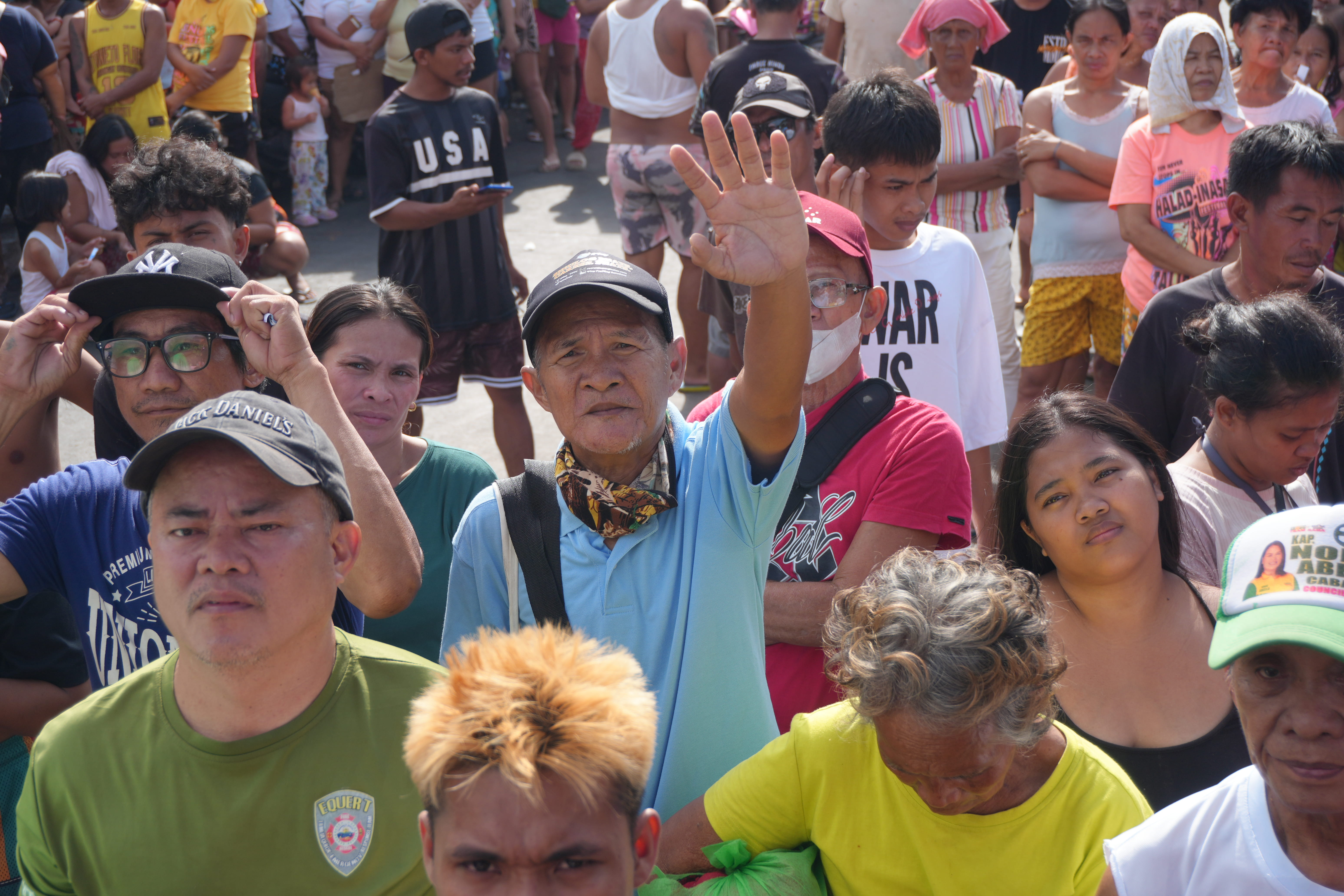 A diverse group of people gathered outdoors with one man in a blue shirt raising his hand.