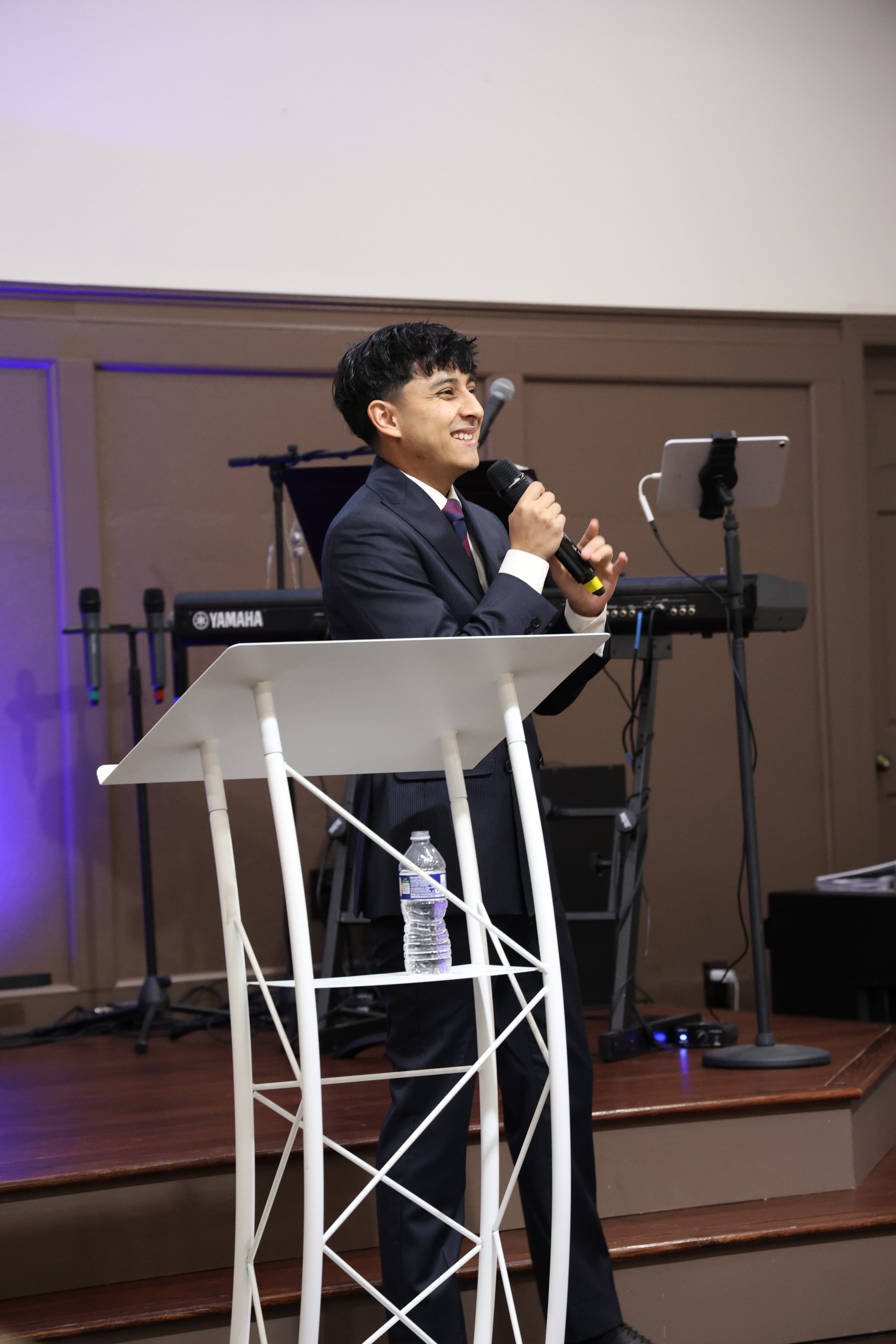 Smiling man in suit speaking into a microphone behind a white podium with a water bottle on it.