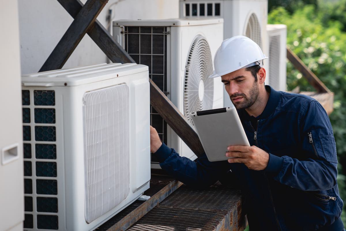 HVAC technician in hard hat inspecting air conditioning unit with tablet