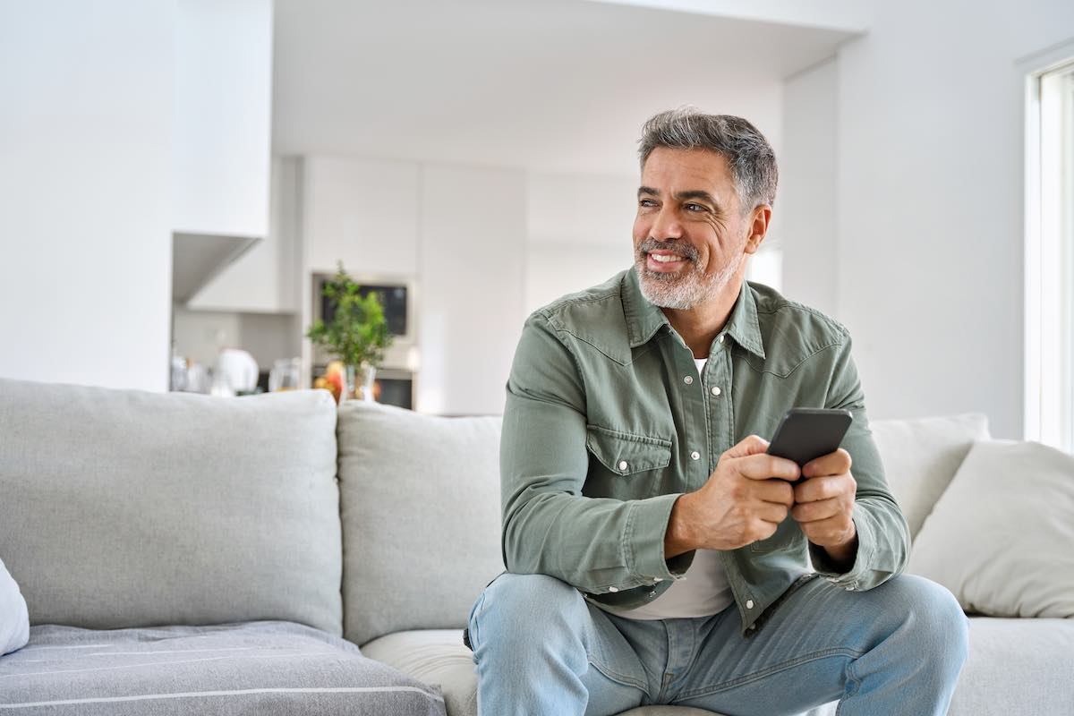 Middle-aged man smiling while holding smartphone on modern living room couch