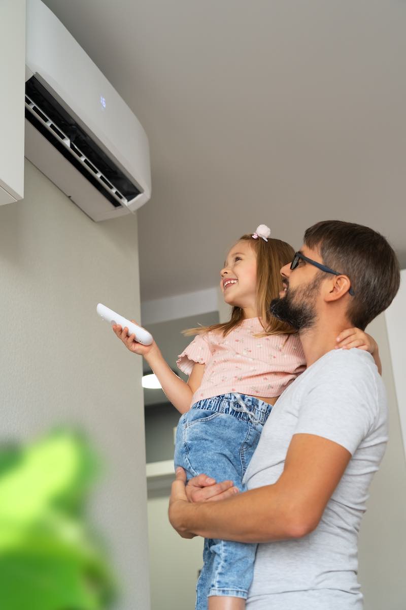 Father holding daughter with remote, adjusting air conditioning unit indoors.