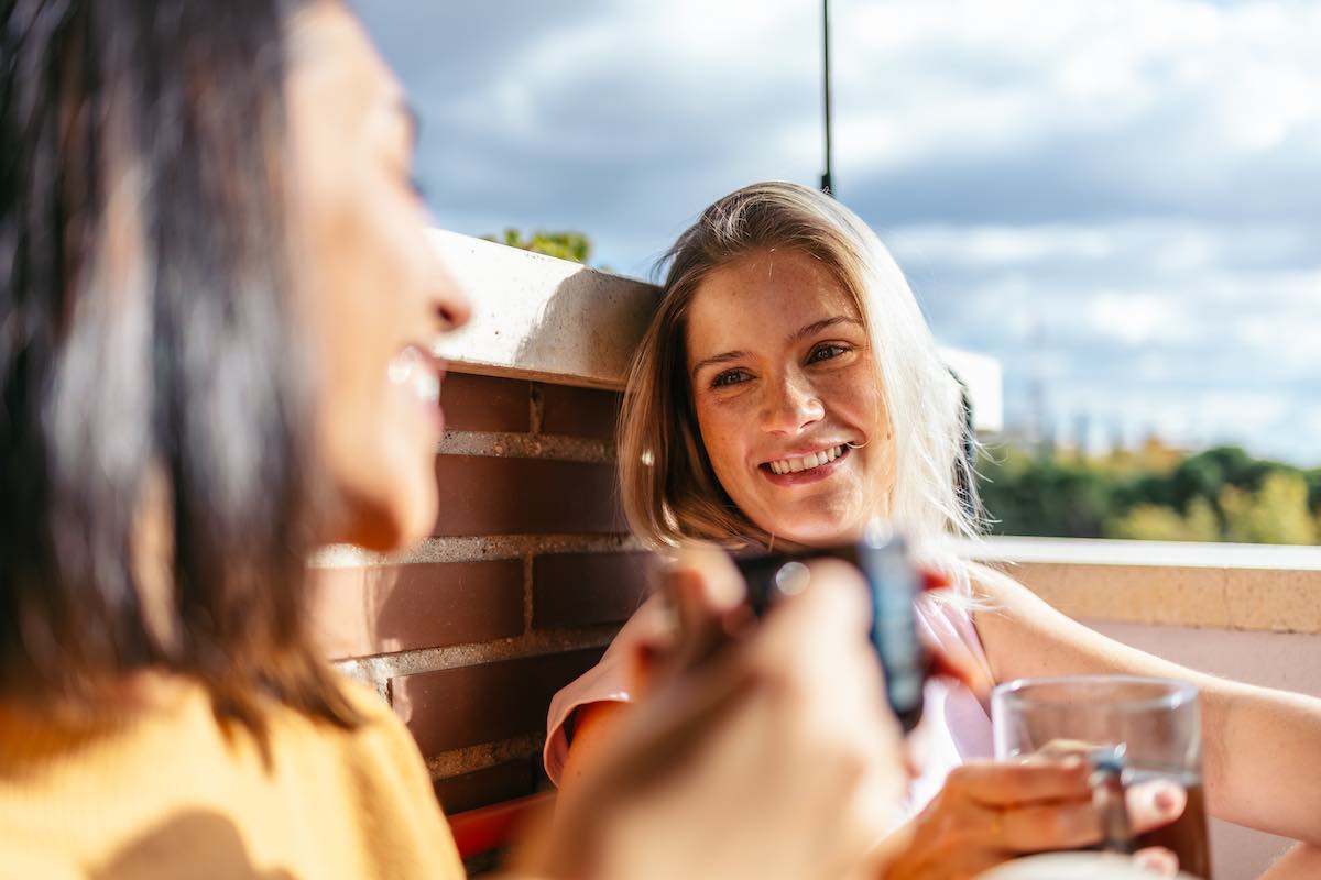 Two young women smiling outdoors on a sunny day, one holding a phone, brick wall background