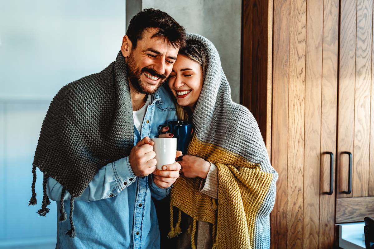 Happy couple wrapped in blankets holding coffee mugs indoors by window