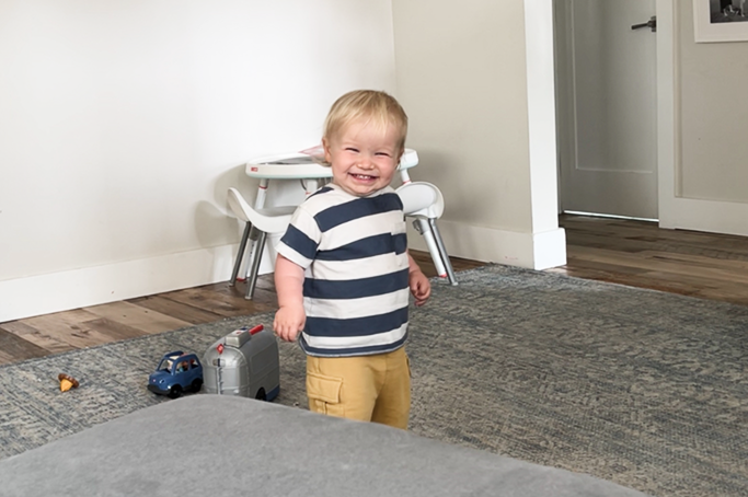Smiling toddler in a striped shirt and yellow pants standing on a gray carpeted floor in a living room with toys and a small white table and chairs in the background.