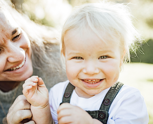 Smiling toddler with light hair wearing overalls, playing with an adult who is smiling beside them.