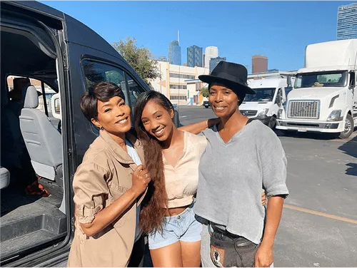 Three smiling women standing next to a black van on what appears to be a film set in a city.
