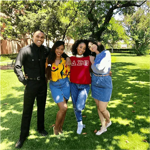Behind-the-scenes shot of four young actors, one in a cop outfit and two women in denim skirts, standing together on a grassy lawn under trees, smiling. 