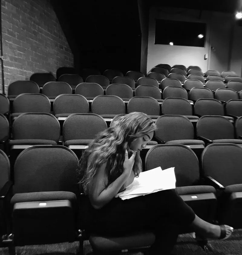 Black and white image of a woman with long wavy hair, sitting in an empty auditorium seat and intently reading a script or book. 