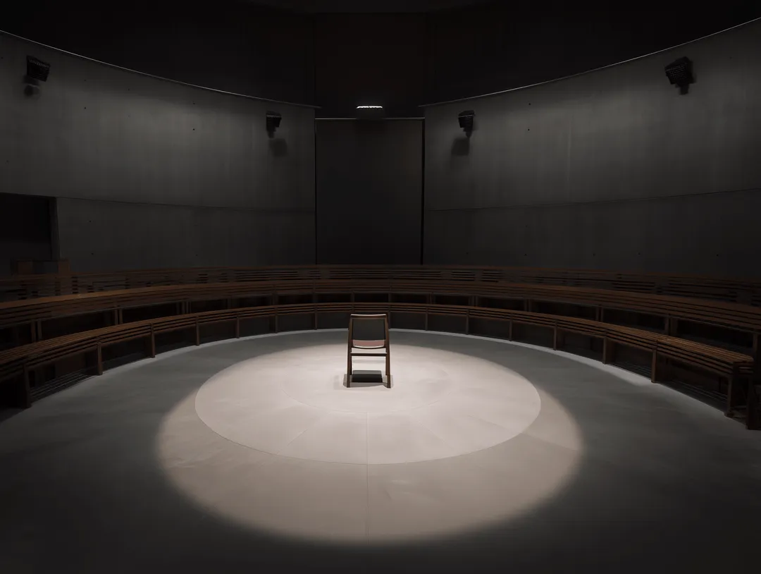 A single wooden chair illuminated by a spotlight in the center or a dark, empty auditorium with tiered seating.