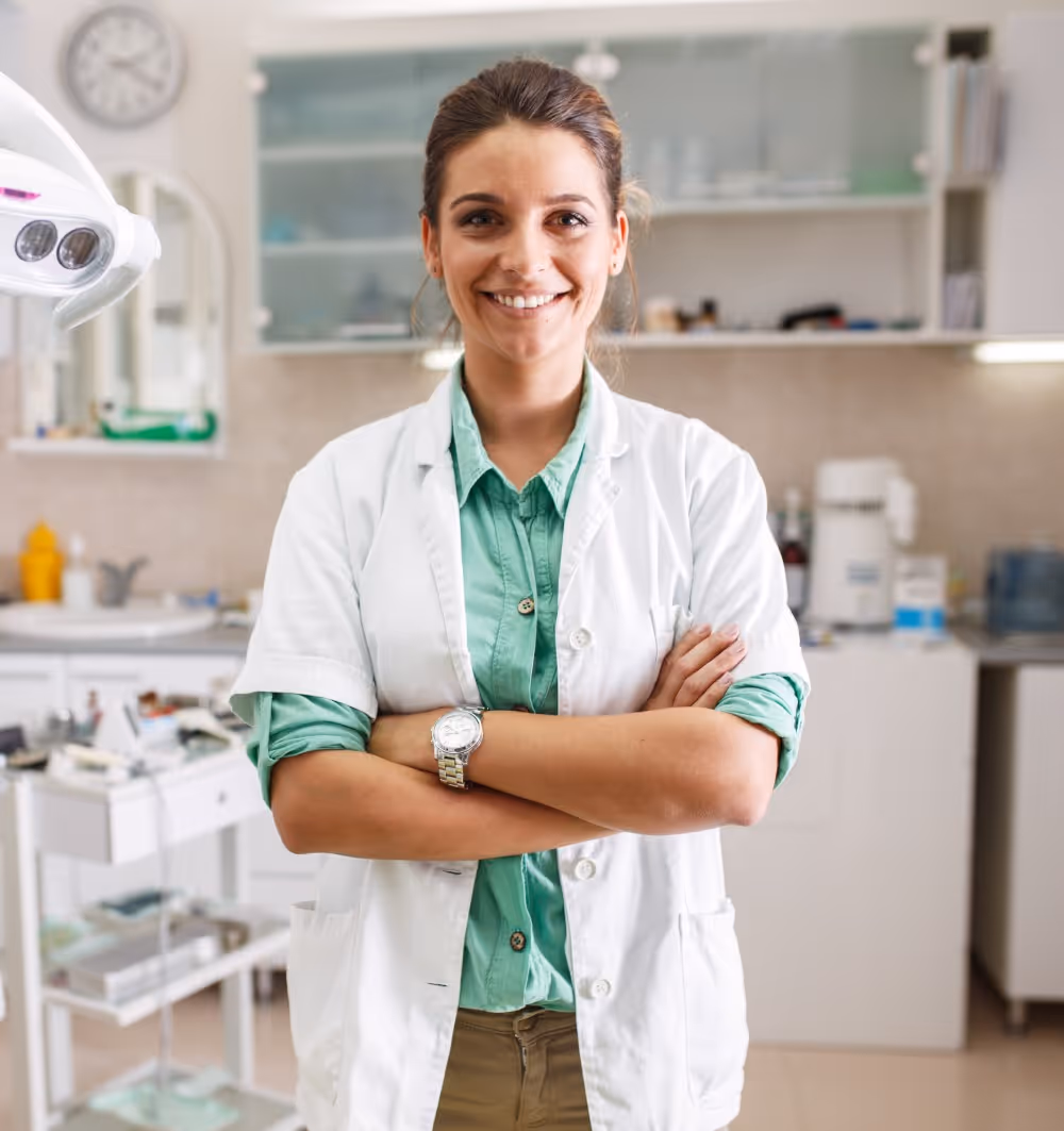 Smiling female dentist in a white coat with arms crossed in a dental clinic.