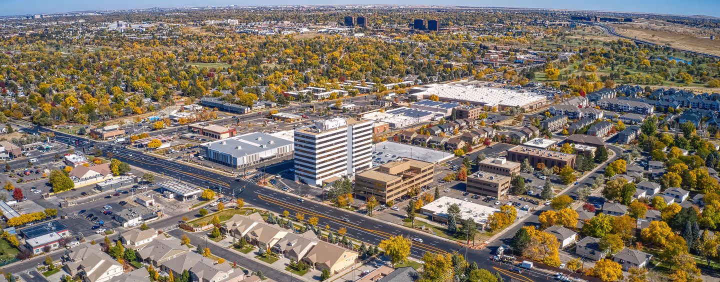 Aerial view of a suburban area with commercial buildings, residential houses, and tree-lined streets showing autumn foliage.