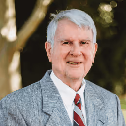 Smiling elderly man with gray hair wearing a gray suit jacket, white shirt, and striped tie standing outdoors.