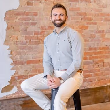 Smiling man with beard sitting on a black stool against a brick wall background.