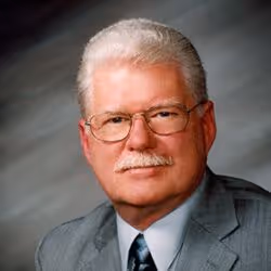 Portrait of an older man with white hair and mustache wearing glasses, a gray suit, white shirt, and dark tie.