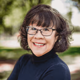 Smiling woman with short dark hair and glasses wearing a black turtleneck outdoors.