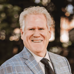 Smiling middle-aged man with curly white hair wearing a gray plaid suit and black tie standing outdoors.