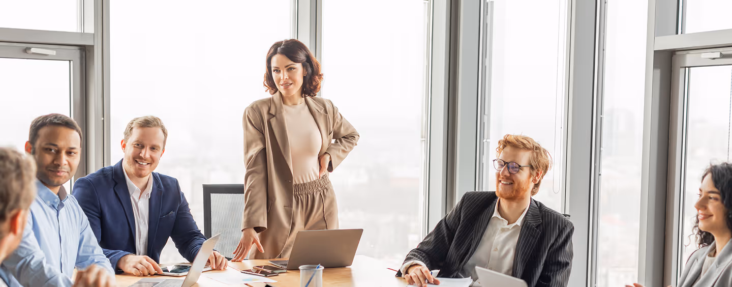 A group of diverse business professionals gathered around a conference table during a meeting with a woman standing and speaking.
