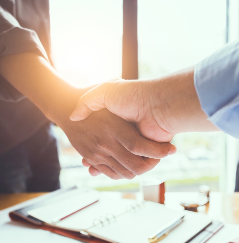 Two people shaking hands over a desk with documents and a pen, illuminated by sunlight through a window.