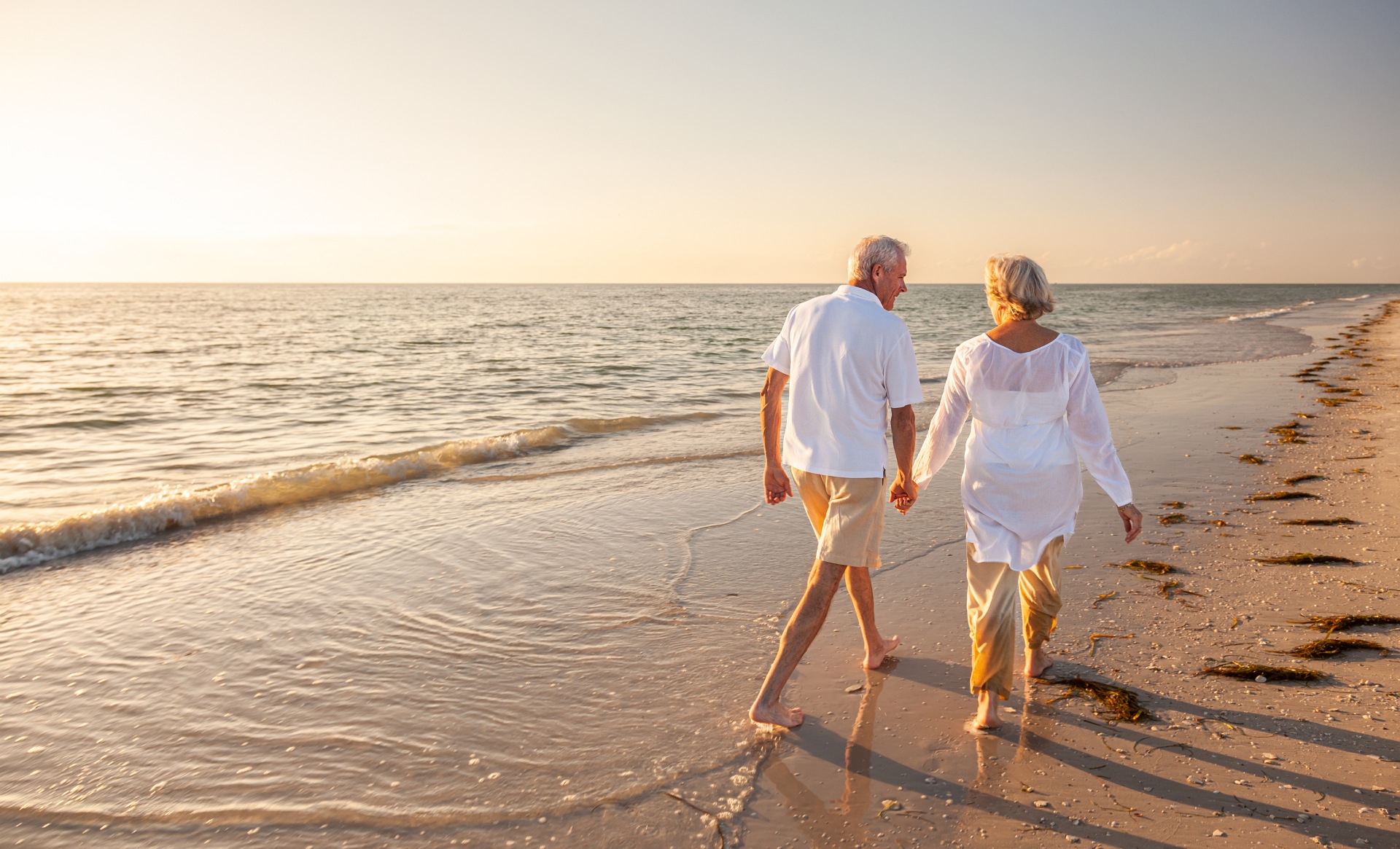 Older couple holding hands and walking barefoot on a sandy beach near gentle waves at sunset.