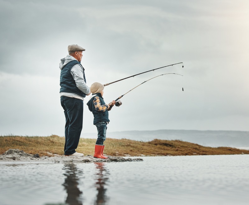 An older man and a child fishing with rods by a calm lake under a cloudy sky.