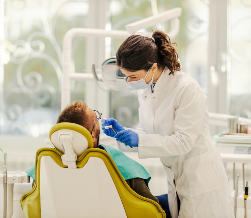 Dentist wearing a mask and gloves examines a male patient seated in a dental chair.